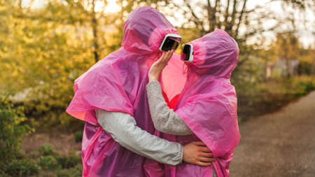 A closeup shot of two people in pink plastic raincoats and VR headset romantically hugging each otherの写真素材