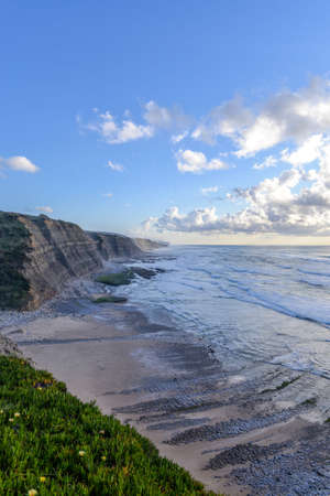A vertical picture of the beach surrounded by the sea and cliffs under the sunlight and a cloudy sky