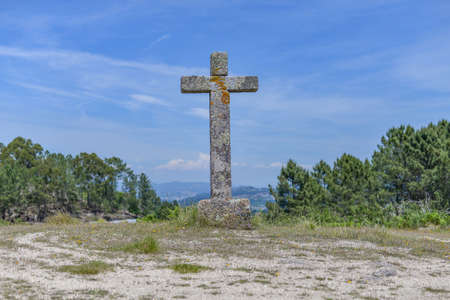 A stone cross covered in mosses surrounded by trees and hills under a blue sky and sunlight