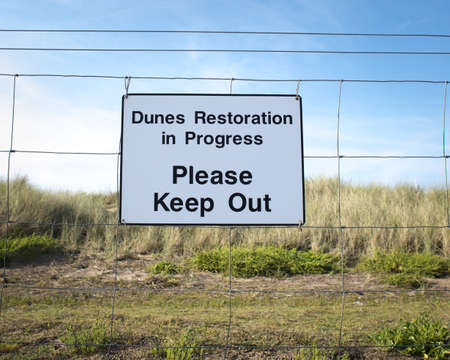 A closeup shot of a sign on a wire fence that says "dunes restoration in progress please keep out"の写真素材