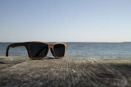 A closeup shot of sunglasses on a wooden surface in front of the waterの写真素材
