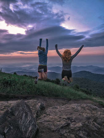 A vertical shot of two females jumping on a cliff under the breathtaking cloudy sky