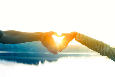 Two hands forming a heart shape with the lake on a blurry background during the sunset