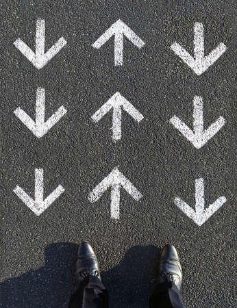 A vertical high angle shot of a male standing in front of arrow signs on the asphalt ground pointing upwards and downwardsの写真素材
