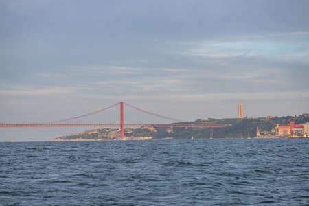 A sea with a red bridge on it surrounded by greenery and buildings under a cloudy sky in Portugal