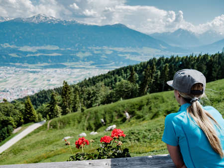 A girl with a blue shirt and a cap surrounded by mountains covered in forests under a cloudy sky