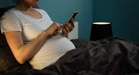 A pregnant woman in a white shirt in her bed holding a phone in her handsの写真素材