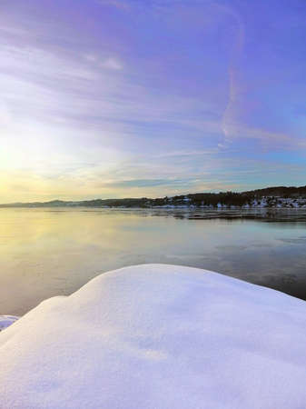 A vertical picture of the snow surrounded by the sea and buildings during the sunset in Norwayの写真素材