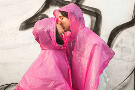 A closeup shot of a male and a female in pink plastic raincoats kissing each otherの写真素材