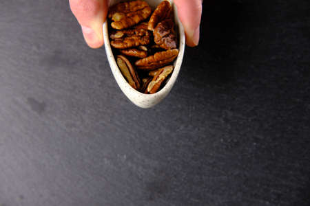Two fingers are holding a small white plate full of brown pecans on a black background
