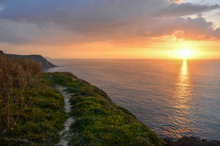 The sea surrounded by cliffs under a cloudy sky with the sun reflecting on the water during the...