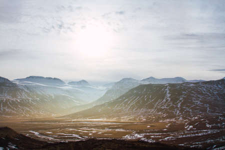 A landscape of hills covered in the grass and snow under a cloudy sky and sunlight in Iceland