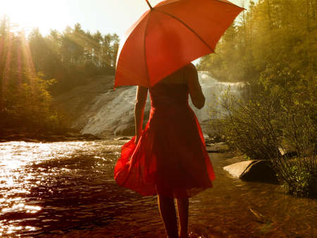 A young female with a red dress holding a red umbrella walking near a waterfallの写真素材