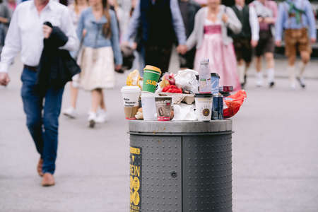 MUNICH, GERMANY - Sep 22, 2019: Overflowing trash can waste bin at world famouse Oktoberfest festival in munich with blurry people in background wearing traditional bavarian trachtのeditorial素材