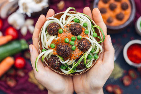 A high angle shot of a person holding a bowl of delicious vegetable meatballs with creamy sauce and zucchini noodles with a blurred backgroundの写真素材