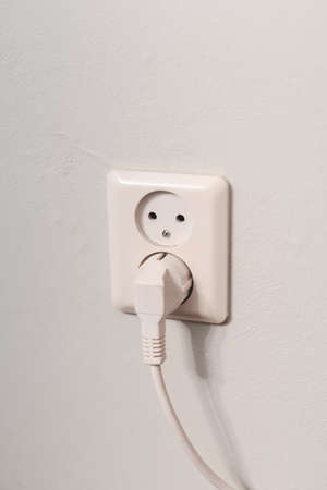 A vertical shot of a wire plugged in the electrical socket on a white wall