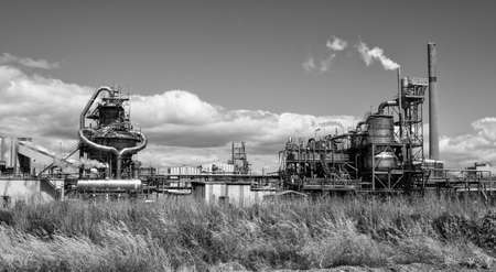 A greyscale of a factory surrounded by the grass under the sunlight and a cloudy sky
