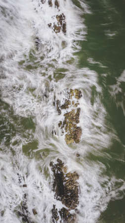 The vertical shot of the ocean waves crashing into the rocks near the beach on a sunny day