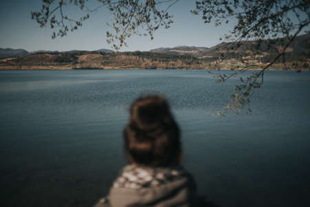 the rear view of a blurry lady in the foreground wearing a bun looking  at a scenic lakeの写真素材
