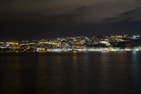 The sea surrounding a city covered in buildings and lights under a cloudy sky during the nightの写真素材