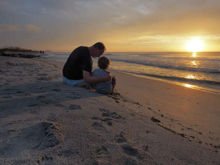 A man hugging his kid while sitting on the beach surrounded by the sea during the sunsetの写真素材