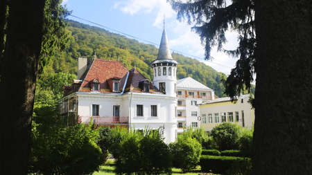 A manor house with white walls and a red roof in a garden surrounded by hills and forests in Romania