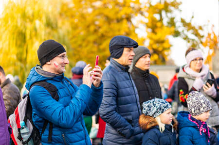 POZNAN, POLAND - Nov 11, 2019: Man with with painted Polish flag on his cheek taking a photo with his smartphone during independence day in the city center.のeditorial素材
