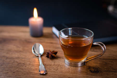 A selective focus shot of star anise tee with a teaspoon, black notebook, eyeglasses, and candle on a wooden tableの写真素材