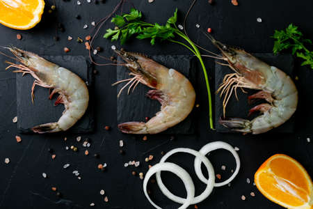 A top view of raw shrimps with onions, herbs, and oranges on a black wooden boardの写真素材