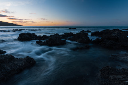 A beautiful scenery of a sea with rocks and hills under a blue an orange sky at sunset