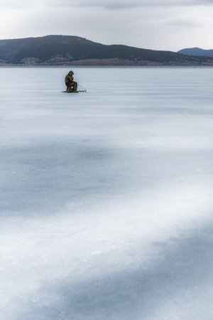 A vertical shot of fisherman fishing in the frozen lake in the mountainsの写真素材