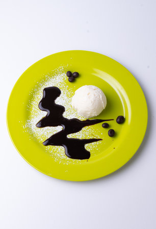 An overhead shot of a scoop of vanilla ice cream on a yellow plate with powdered sugar, chocolate syrupの写真素材