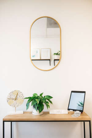 A vertical shot of clock design, house plant and a tablet in a table with a mirror on a white wallの写真素材