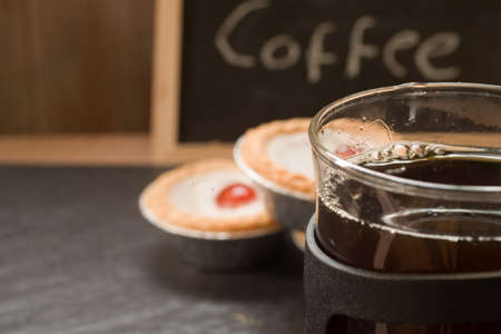 A closeup shot of coffee in a glass mug with a blurry coffee word and tartlets in the background