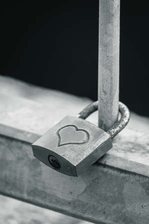 A closeup grayscale shot of a metal padlock with a heart hanging on a fenceの写真素材
