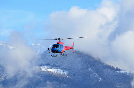 A blue and red helicopter in the air above the hills covered in greenery at daytimeの写真素材