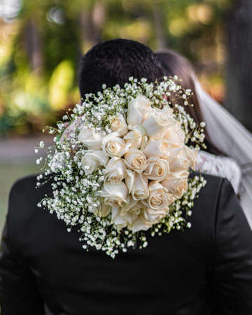 A beautiful shot of a bride holding a beautiful bouquet of roses while hugging her groom with a...