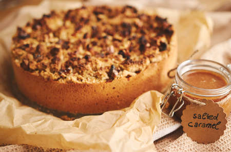 A selective focus shot of a round apple pie topped with crumble and nuts, on a towel, and a jar of...