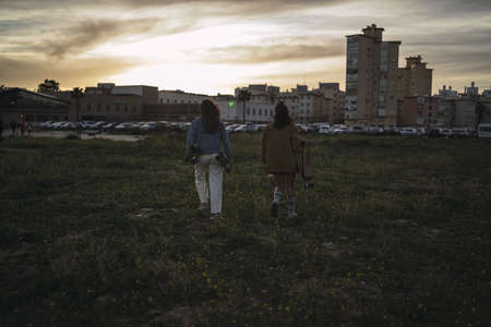 The two young females with skateboards walking through the green field at sunsetの写真素材
