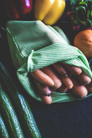 A vertical closeup shot of carrots in a green bag and other vegetables on a dark surfaceの写真素材