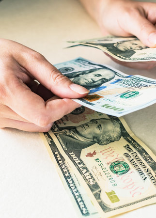 A closeup of a woman's hands counting dollar bills, on a white desk.