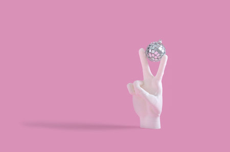 A human hand showing the v sign and holding a disco ball on a pastel pink background
