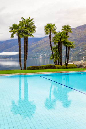A swimming pool surrounded by palm trees and an Alpine Lake in Ascona in Switzerlandのeditorial素材