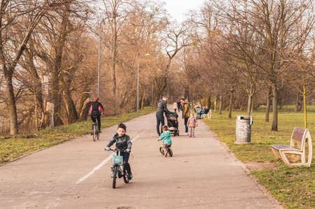 POZNAN, POLAND - Mar 18, 2020: Parents and kids with bikes and by feet on a footpath along trees at a park.のeditorial素材