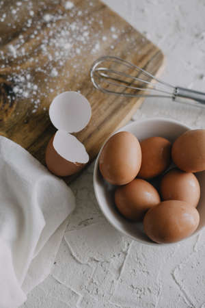 A vertical shot of a bowl of eggs and a silver whisk next to a chopping board on a white surface