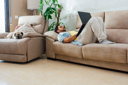 A young man resting on a sofa at home and using a laptop with his dog beside himの写真素材