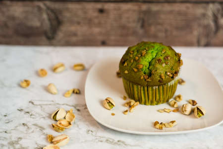 Single Pistachio Muffin on white plate against a wood backdrop with white counter surrounded by...