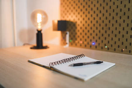 A selective focus shot of an open notebook with a black pen and a lamp on a wooden deskの写真素材