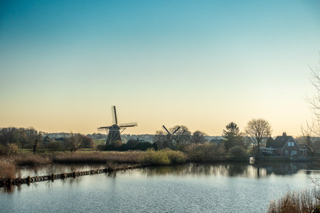 A beautiful shot of the windmill near the river surrounded by trees and houses