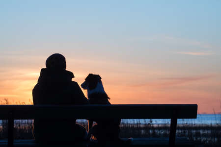 Silhouette portrait of woman and her dog sitting on the bench in the beach and enjoying the setting sun on the seaの写真素材